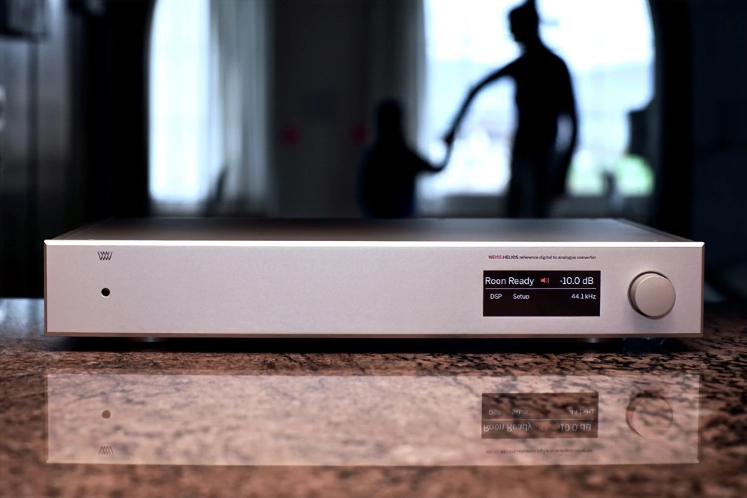 Un DAC Weiss Helios haut de gamme affichant "Roon Ready" et "-10.0dB" sur un comptoir en pierre brillant, avec une silhouette floue de deux personnes en arrière-plan (par 1877.audio)
