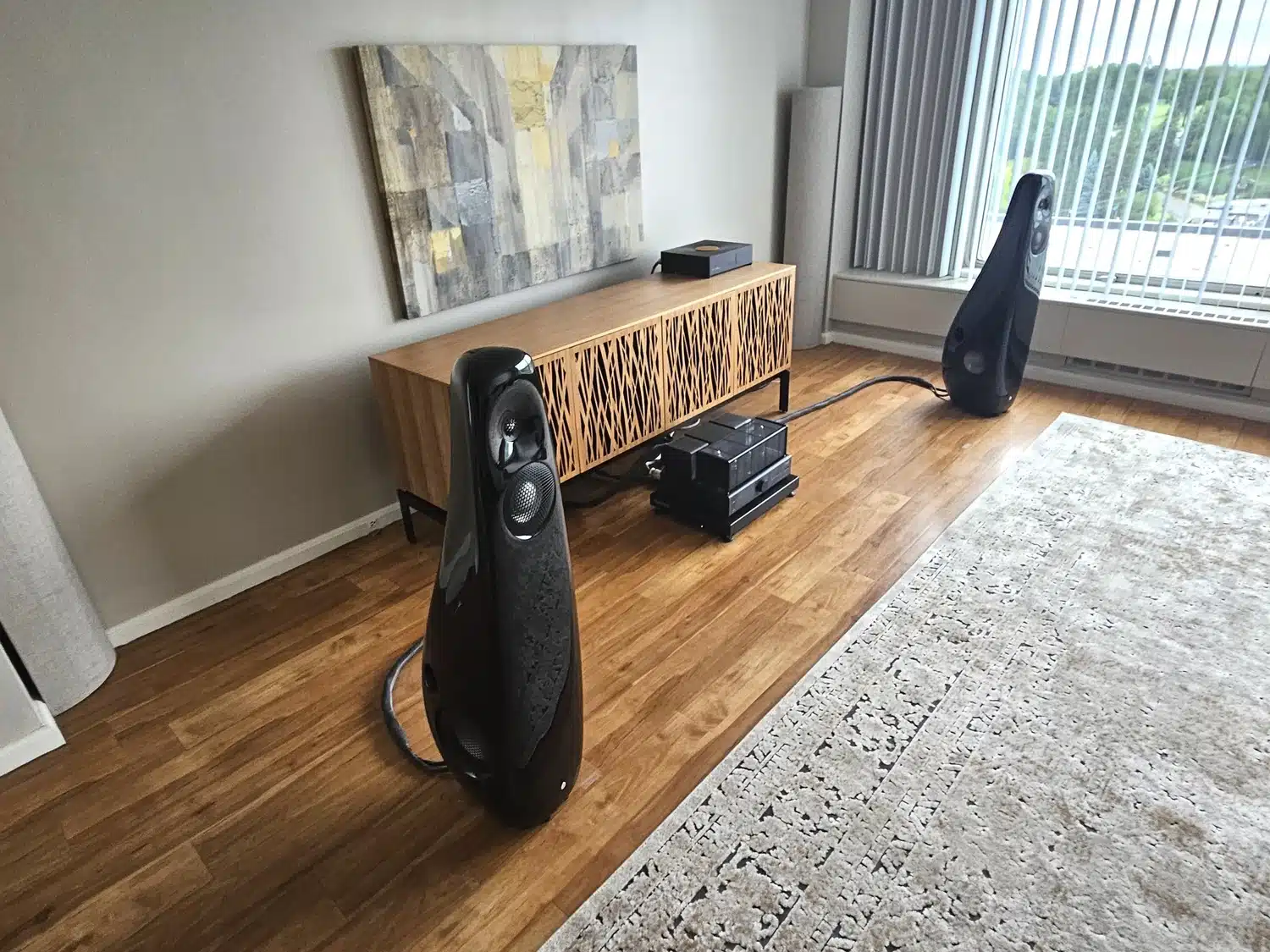 Un salon moderne avec un parquet en bois, une grande peinture abstraite au-dessus d'une console en bois, flanquée de deux enceintes noires Vivid Audio Kaya 45 (par 1877.audio)
