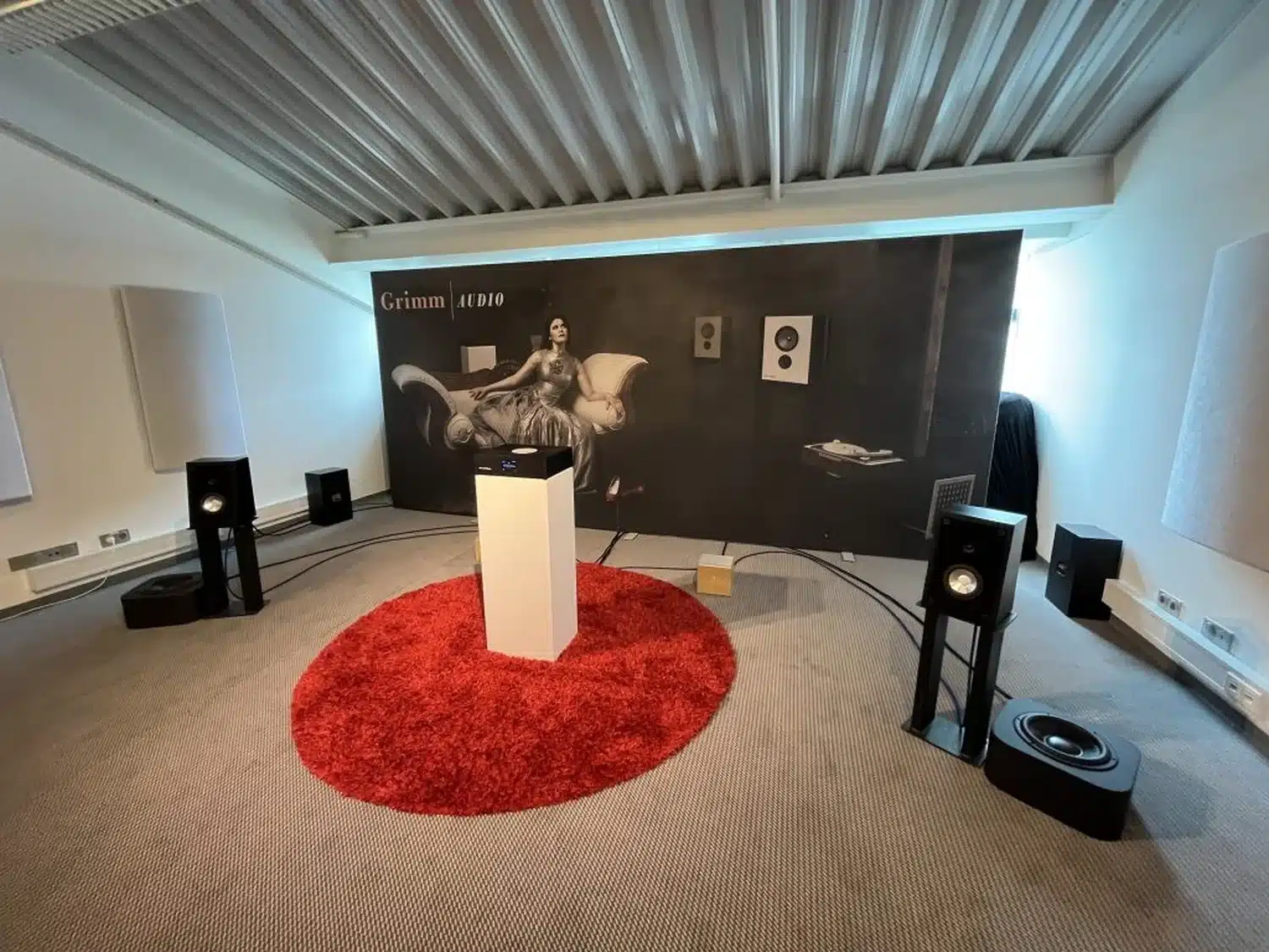 Salle d'écoute au Salon de Munich 2023 avec tapis rouge, deux enceintes noires avec caisson Grimm Audio SB1, une fresque murale pour présenter le MU2 en avant-première (par 1877.audio)