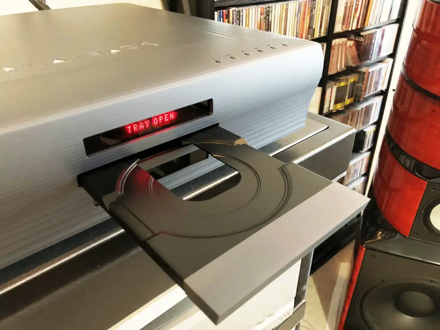 Un lecteur de CD/SACD Playback Designs MPS-8 avec son plateau ouvert vide configuré pour la lecture avec en fond d'image une étagère remplie de pochettes de CD (par 1877.audio)