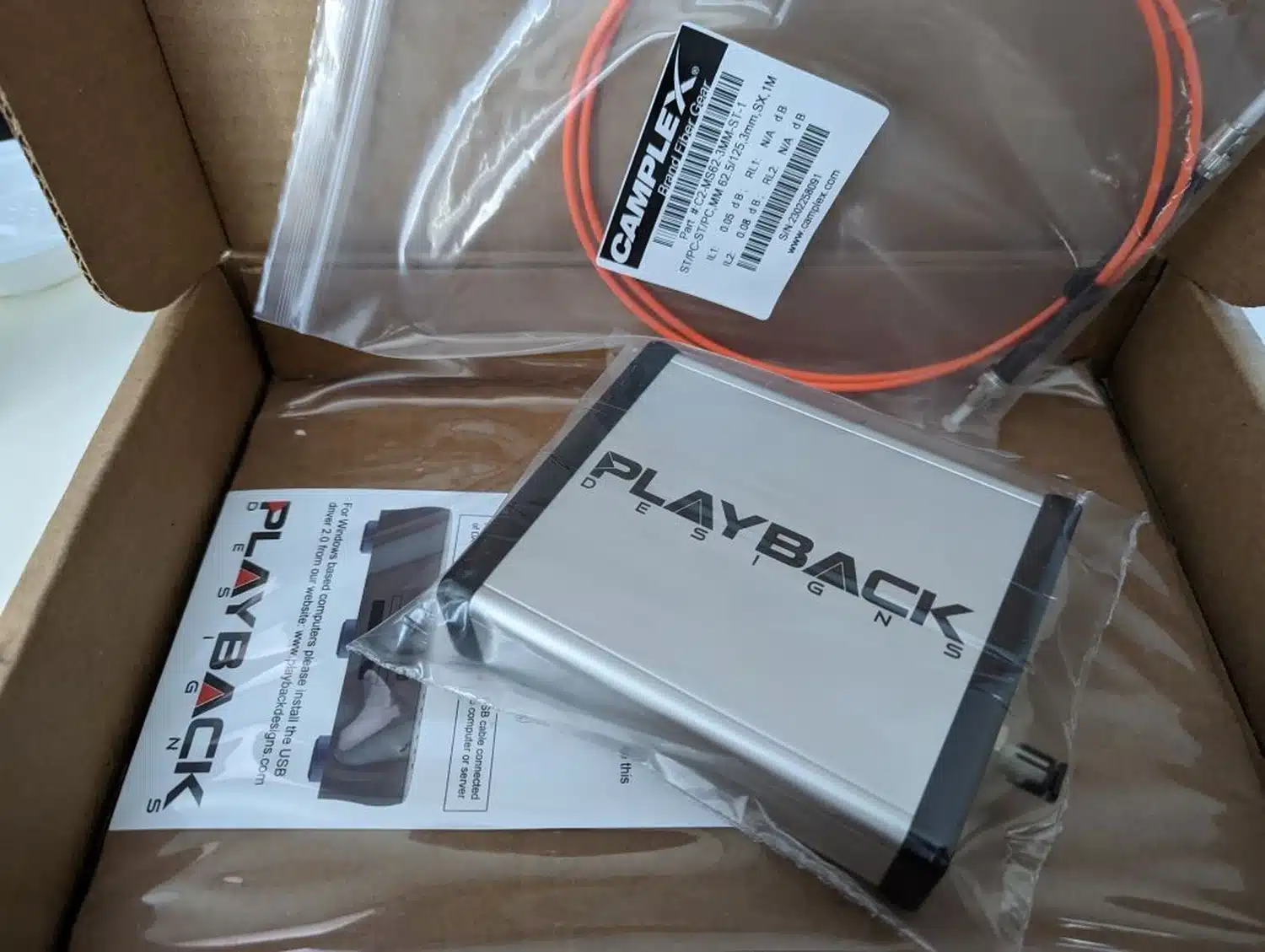 Boîte en carton ouverte contenant une interface USB-X4 de Playback Designs ainsi que son câble fibre optique orange, tous deux scellés dans leur sac en plastique transparent (par 1877.audio)