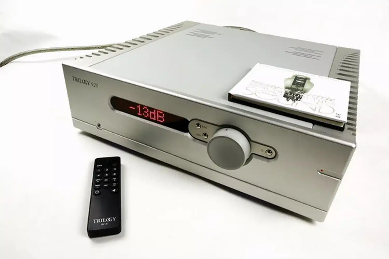Un amplificateur hybride intégré Trilogy Audio 925 avec son affichage numérique rouge indiquant "-13db", accompagné à côté de sa télécommande noire et d'une pochette de CD sur le dessus (par 1877.audio)