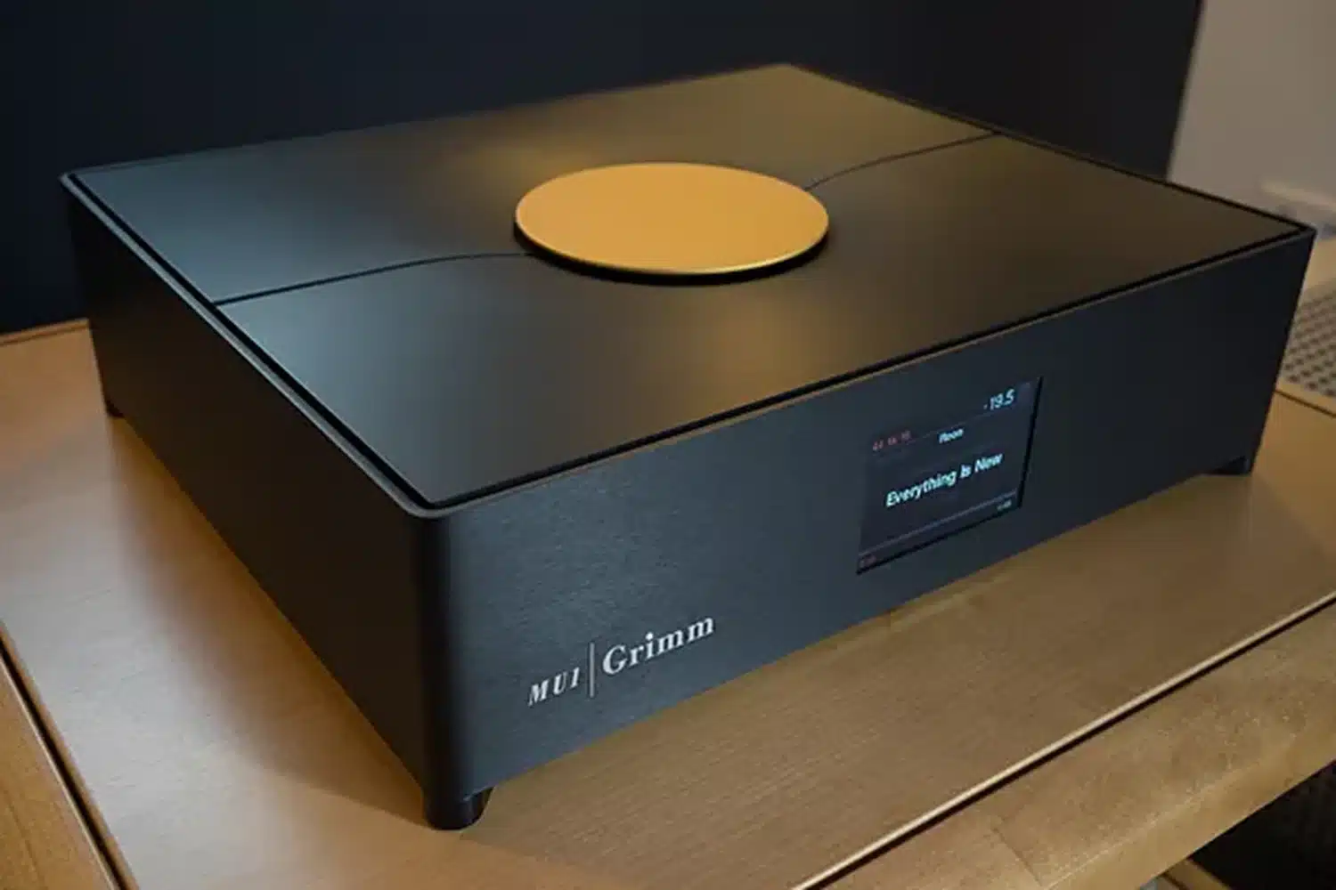 L'appareil de streaming Grimm Audio MU1 noir avec son disque de commande doré sur la face postérieure affichant des informations sur le morceau en cours de lecture sur son écran posé sur une plateforme en bois (par 1877.audio)