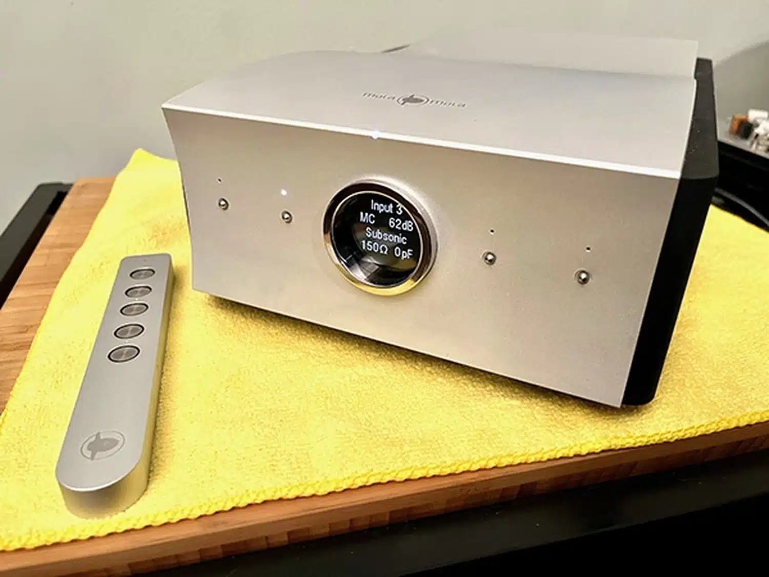 Un amplificateur phono Mola-Mola Lupe haut de gamme doté d'un affichage numérique rond indiquant que la troisième entrée est sélectionnée (avec les paramètres MC, 62dB, Subsonic, 150Ohms et 0pF) et de sa télécommande, le tout posé sur un tissu jaune (par 1877.audio)