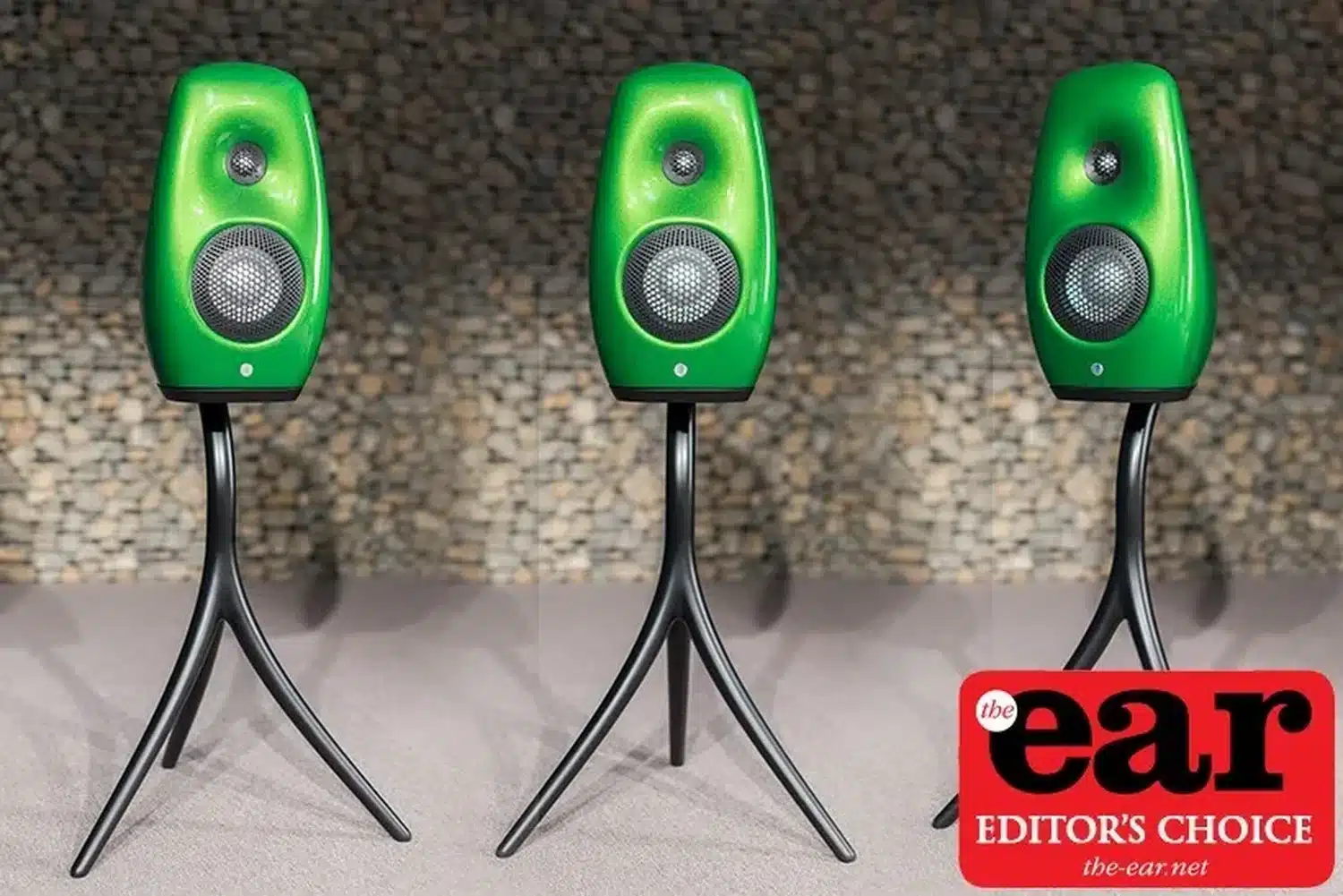 Trois enceintes vertes haut de gamme Vivid Audio Kaya S12 sur leur trépied spécifique se dressent avec des angles différents sur un fond gris en galets avec une étiquette « Editor's Choice » de The Ear (par 1877.audio)