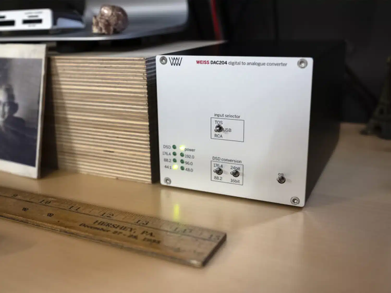Le convertisseur numérique-analogique Weiss DAC204 est posé sur un bureau à côté d'une règle en bois et d'un cadre photo flou en arrière-plan. Le DAC est alimenté et fonctionne d'après ses LED en 44.1kHz (par 1877.audio)